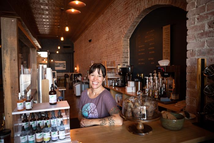 Artisan Mark owner Elayne Woods Jones stands behind the coffeeshop's counter. Behind her, 很长一段, exposed brick wall extends to seating at the back of the shop, with warm lights overhead.